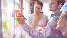 Eine junger Mann schreibt auf einen kleinen bunten Notizzettel, der an einer Fensterscheibe neben vielen weiteren Zetteln hängt, ein Kollege und eine Kollegin schauen zu. Der Raum, in dem sie stehen, ist lichtdurchflutet.