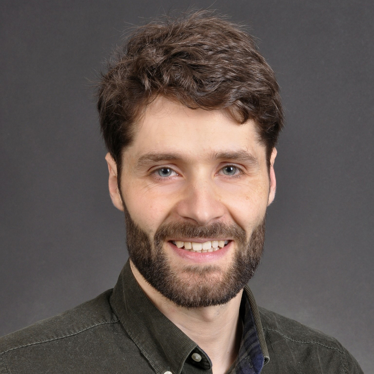 Ein Mann mit dunkelbraunem Haar und Bart l&auml;chelt in die Kamera. Er tr&auml;gt ein dunkelbraunes Hemd. Im Hintergrund ist eine graue Fl&auml;che.