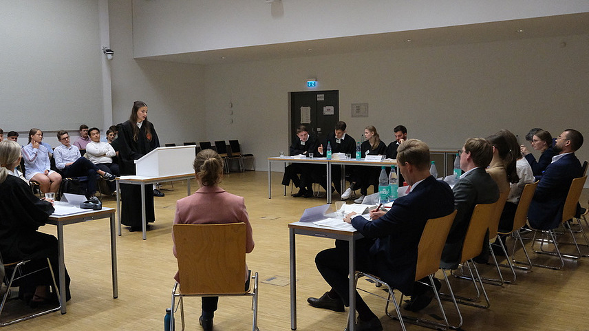 Eine Vortragende in schwarzer Robe vor einer simulierten Richterbank mit den jeweiligen Teams auf der rechten und linken Seite an Tischen sitzend und Publikum im Hintergrund