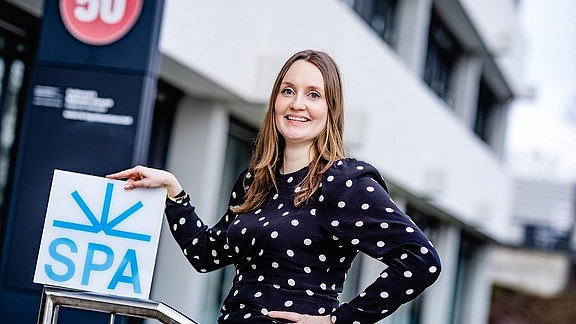 A young woman in a polka-dotted dress is standing in front of a building and presenting the Spa logo.