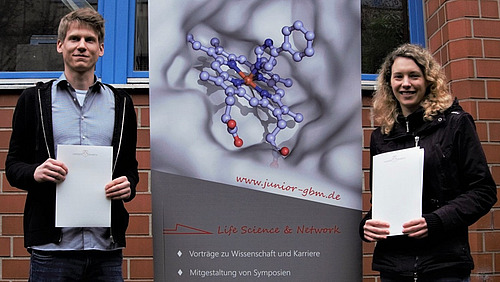 A young man and a young woman are standing in front of a building, smiling and holding certificates in their hands.