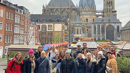 Eine Gruppe von Personen steht dicht beieinander im Vordergrund und schaut in Richtung Kamera. Einige tragen Jacken, M&uuml;tzen oder Schals.  Im Hintergrund sind mehrere Geb&auml;ude zu sehen, darunter H&auml;user mit unterschiedlichen Fassaden sowie ein gro&szlig;es Bauwerk mit vielen Fenstern, T&uuml;rmen und einem weitl&auml;ufigen Dach.  Zwischen Vorder- und Hintergrund befinden sich Marktst&auml;nde mit farbigen D&auml;chern. Der Himmel ist grau.