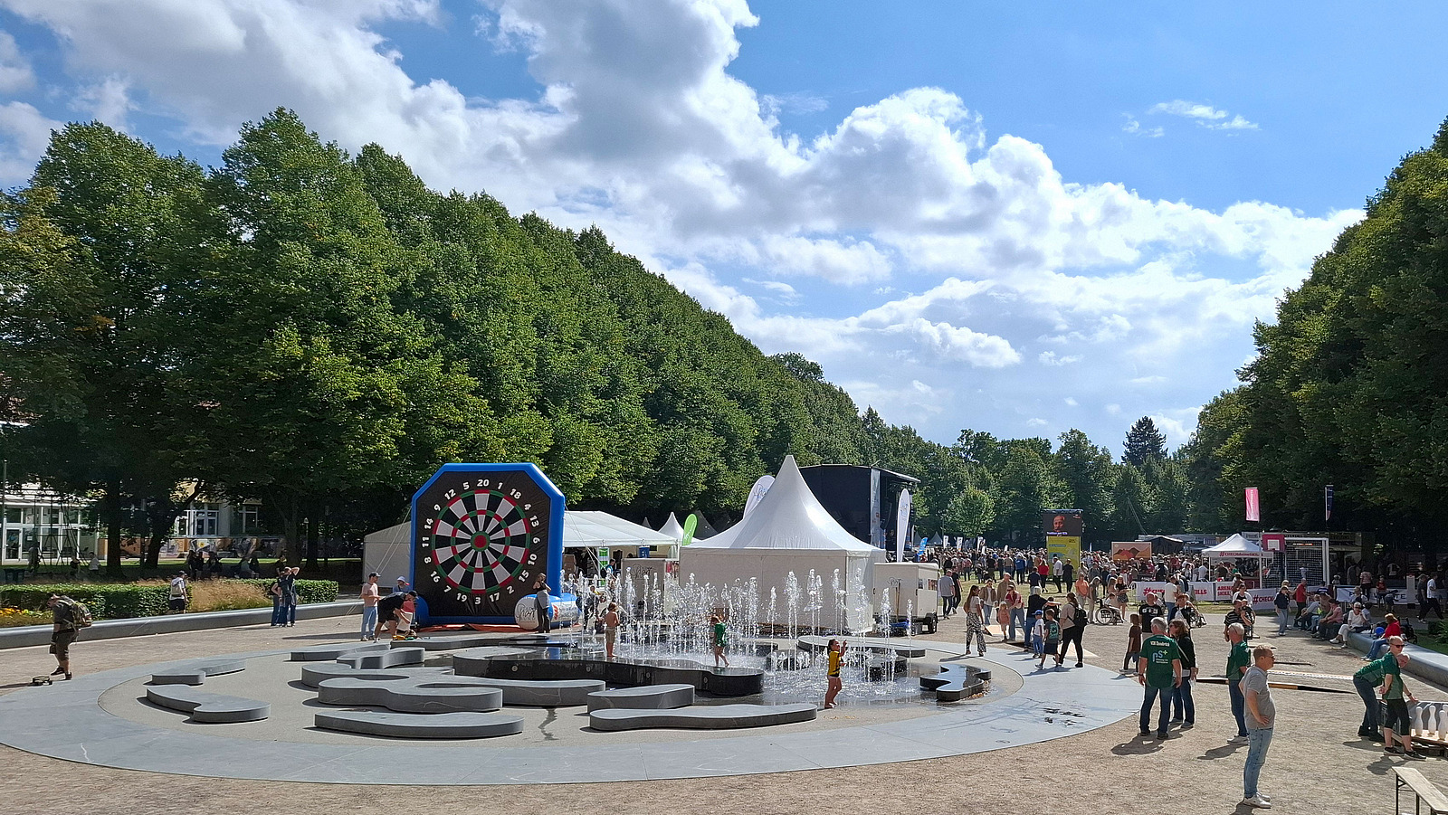 Dartboard and pavilions in a square with a fountain and trees in the background.