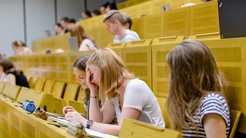 Zwei Studentinnen im Vordergund lachen gemeinsam. Um sie herum sitzen weitere Studierende im Hörsaal auf gelben Sitzbänken.