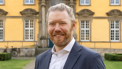 Ein Mann in formeller Kleidung steht lächelnd vor einem historischen Gebäude.