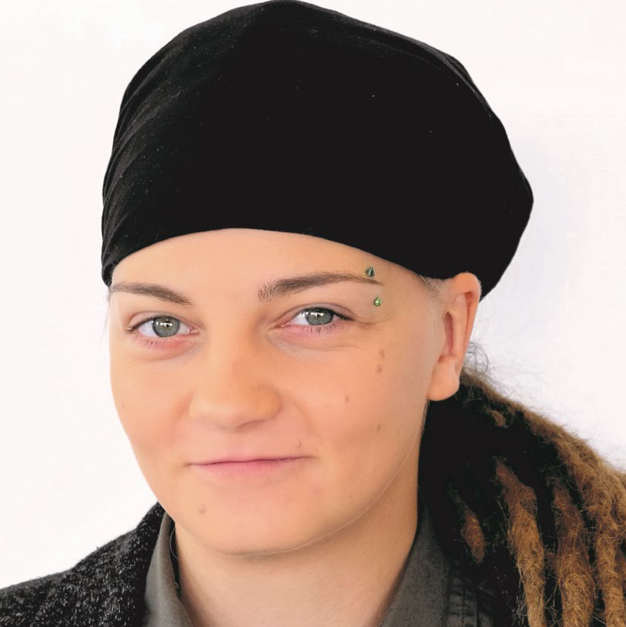 Eine Person mit hellen Augen und Dreadlocks, die unter einem schwarzen Kopftuch hervorschauen, blickt leicht lächelnd in die Kamera. Sie trägt ein dunkles Hemd und eine Strickjacke. 