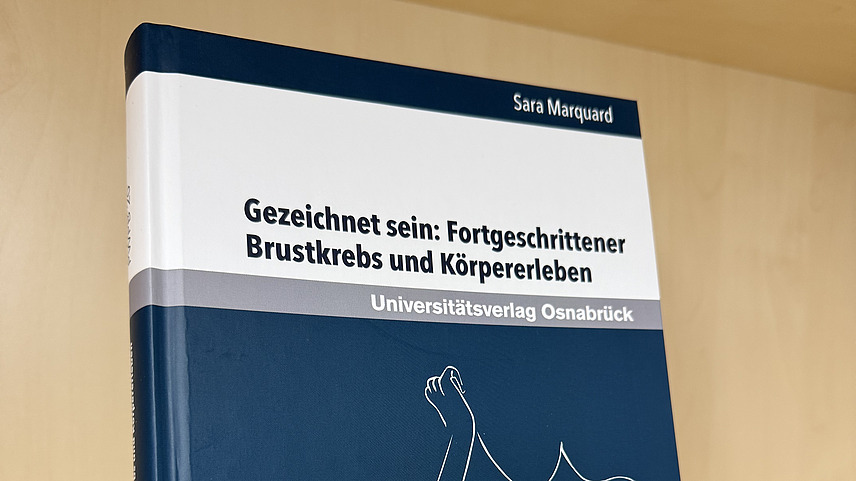 Ein Fachbuch von Sara Marquard mit dem Titel "Gezeichnet sein: Fortgeschrittener Brustkrebs und K&ouml;rpererleben." steht in einem Regal.