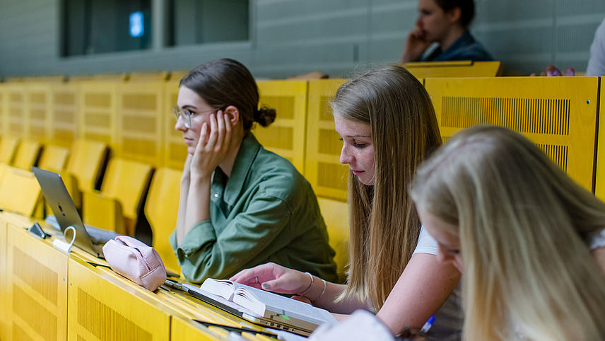 Side view of three female students sitting on a yellow bench in a lecture hall. One is looking forward with concentration, one is reading something in a book in front of her, one is taking notes on a paper pad.
