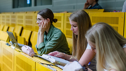 Side view of three female students sitting on a yellow bench in a lecture hall. One is looking forward with concentration, one is reading something in a book in front of her, one is taking notes on a paper pad.