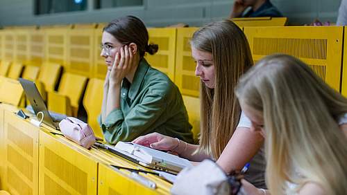 Side view of three female students sitting on a yellow bench in a lecture hall. One is looking forward with concentration, one is reading something in a book in front of her, one is taking notes on a paper pad.