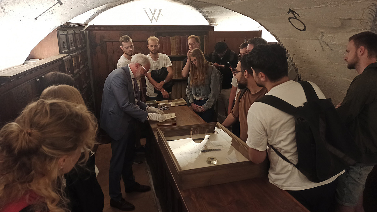 Das Bild zeigt eine Gruppe Menschen in einem Gewölbekeller. An den Wänden stehen braune Schränke. In der Mitte steht ein Mann im grauen Anzug und weißen Handschuhen über einem alten Buch.