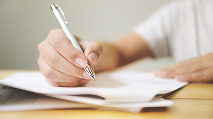 A person sits at a table and writes on a pile of paper with a pen.