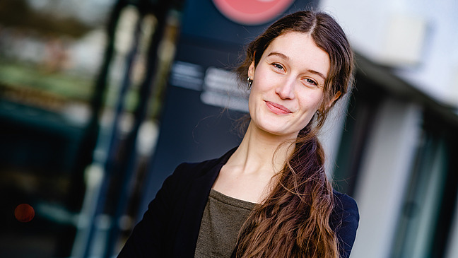 Portrait von einer jungen Frau vor einem Gebäude. Sie trägt einen schwarzen Blazer und lächelt in die Kamera. Ihr langes braunes Haar fällt ihr über die Schulter.
