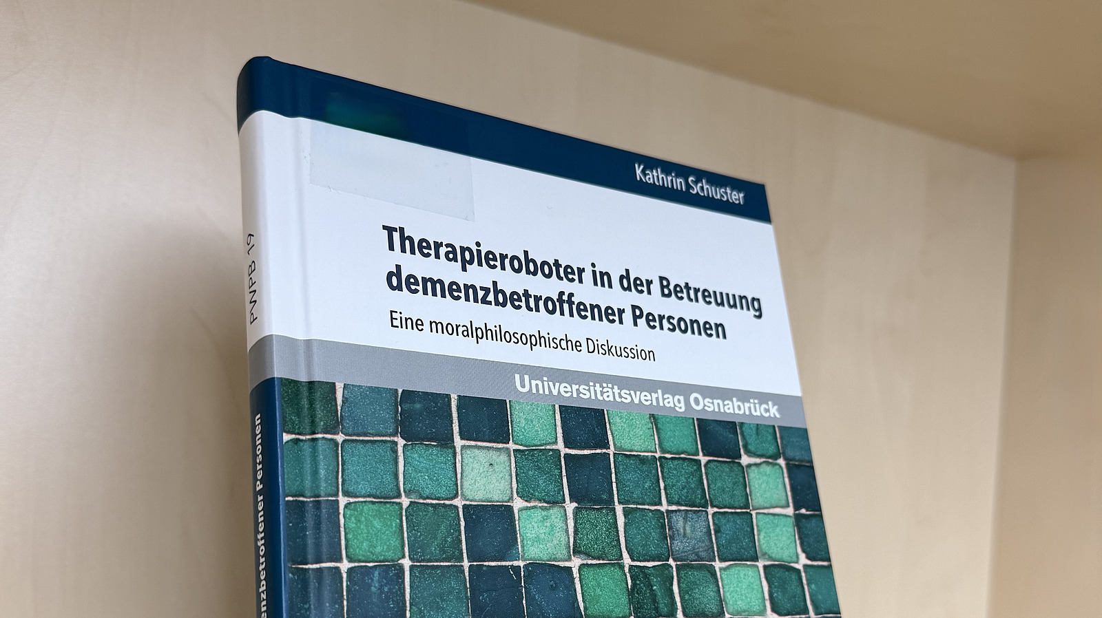 Ein Fachbuch von Kathrin Schuster mit dem Titel "Therapieroboter in der Betreuung demenzbetroffener Personen. Eine moralphilosophische Diskussion" steht in einem Regal.
