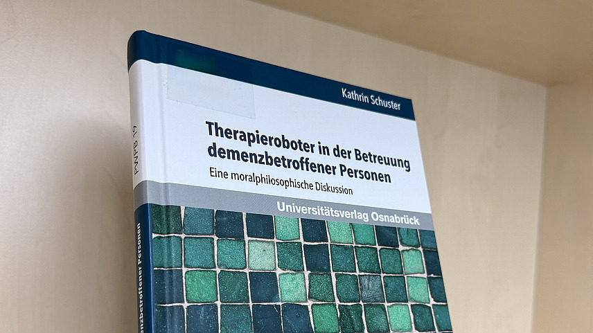 Ein Fachbuch von Kathrin Schuster mit dem Titel "Therapieroboter in der Betreuung demenzbetroffener Personen. Eine moralphilosophische Diskussion" steht in einem Regal.