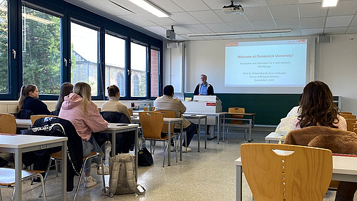 Studierende sitzen in einem Seminarraum, vorn steht Roland Brandt. Hinter ihm an die Wand projiziert steht "Welcome at Osnabr&uuml;ck University!"