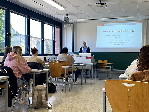 Studierende sitzen in einem Seminarraum, vorn steht Roland Brandt. Hinter ihm an die Wand projiziert steht "Welcome at Osnabr&uuml;ck University!"