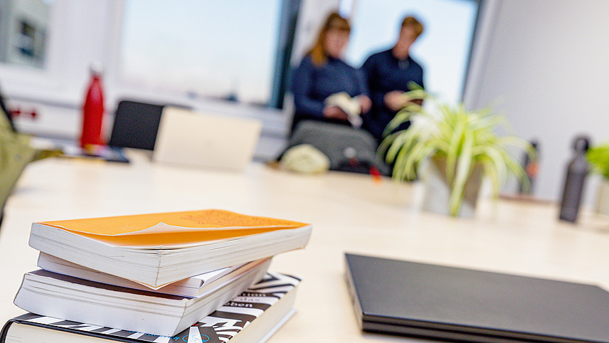 Auf einem Tisch liegen verschiedene B&uuml;cher und ein Computer, die auf ein Projekt oder eine gemeinsame Arbeit hindeuten. Der Hintergrund ist unscharf, zwei Personen stehen an einem Fenster und sind an einer T&auml;tigkeit beteiligt.  