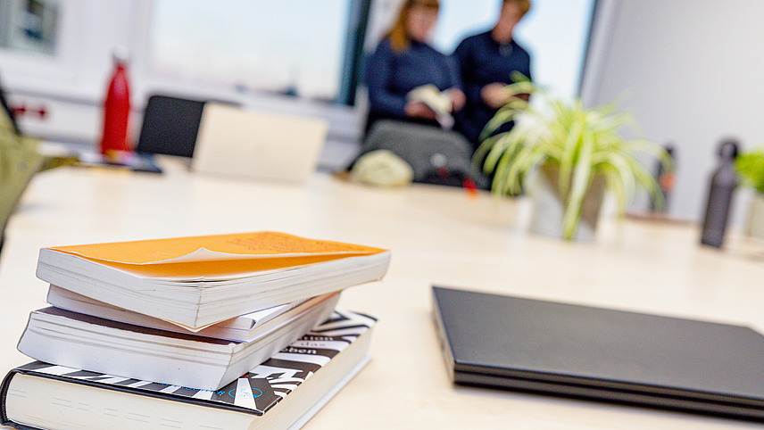 Various books and a computer lie on a table, indicating a project or joint work. The background is blurred, two people are standing at a window and are involved in an activity.  