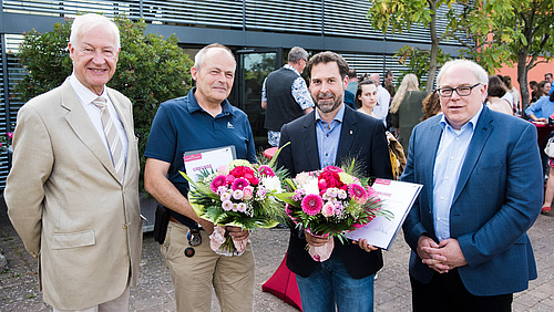 Four men smile at the camera, the two in the middle holding flowers and certificates.
