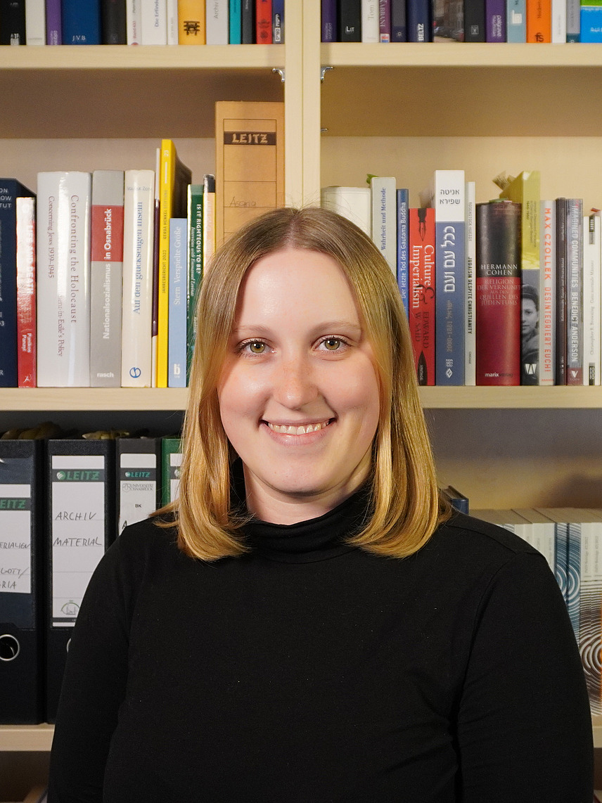 Eine junge Frau mit schulterlangem, blondem Haar lächelt freundlich in die Kamera. Sie trägt ein schwarzes Oberteil und steht vor einem gut gefüllten Bücherregal. Die Regale sind mit Büchern in verschiedenen Größen und Farben sowie einigen beschrifteten Aktenordnern bestückt.