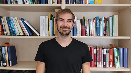 Ein junger Mann mit braunen Haaren und Bart lächelt in die Kamera, vor einem vollgepackten weißen Bücherregal. Die Bücher sind in verschiedenen Farben und Größen angeordnet.