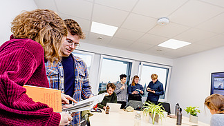 The picture shows a bright room in which several people are present. In the foreground, two people are intensively engaged with a book. In the background, other people can be seen at a table and in front of the windows, standing together as a team and busy with a book and computer. 