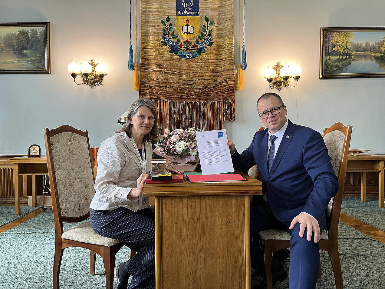 A woman and a man sit at a table and hold a certificate up to the camera. 