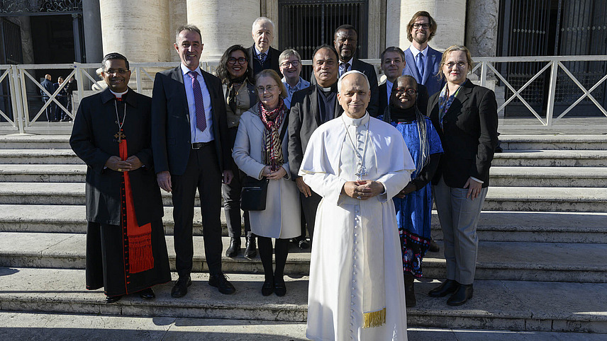 Papst Leo XIV. steht einige Schritte vor dem Komitee. Die Gruppe steht auf einer Au&szlig;entreppe und schaut in die Kamera.