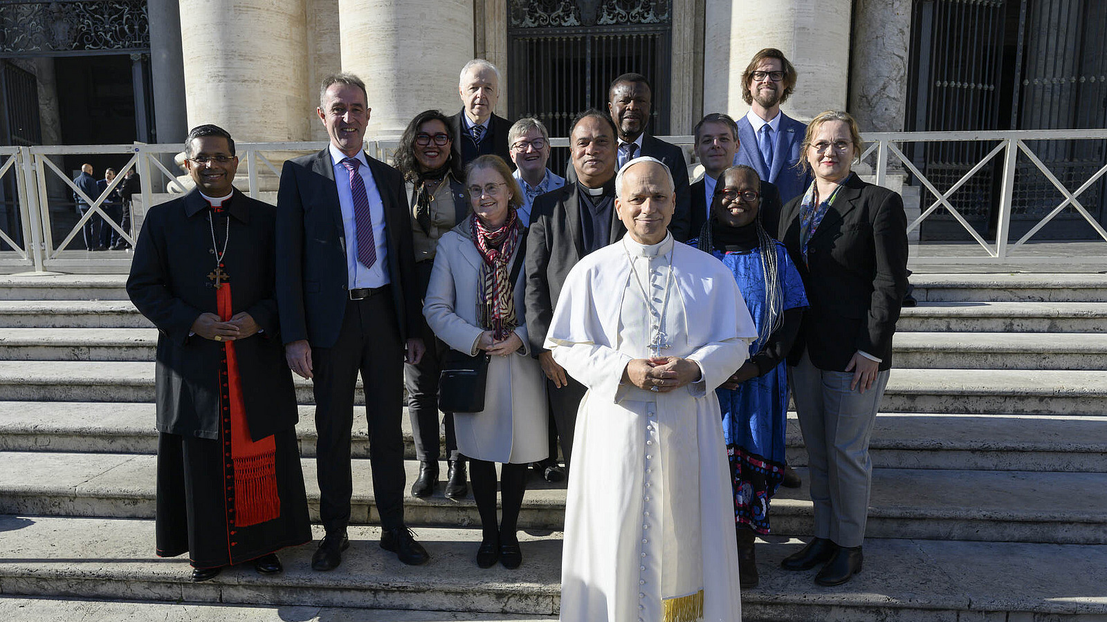 Papst Leo XIV. steht einige Schritte vor dem Komitee. Die Gruppe steht auf einer Au&szlig;entreppe und schaut in die Kamera.
