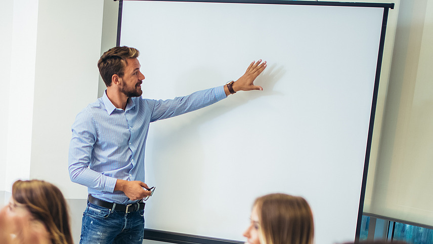 Eine Berufsschulklasse. Der Lehrer steht vorne und zeigt etwas an der Tafel.