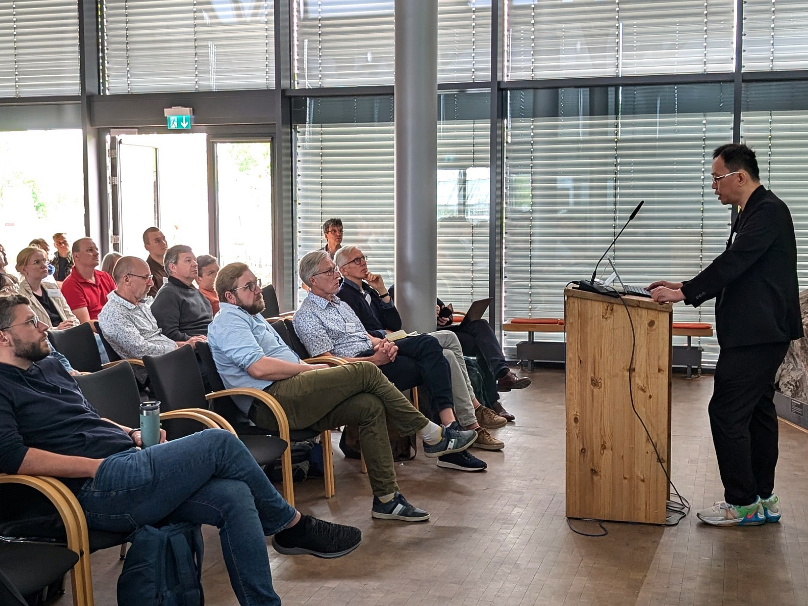 A speaker stands at a lectern, with people sitting on chairs in front of him.