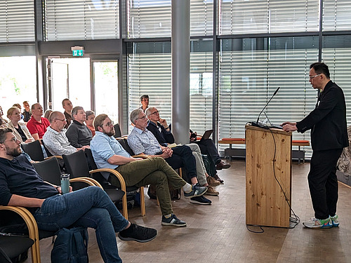 A speaker stands at a lectern, with people sitting on chairs in front of him.