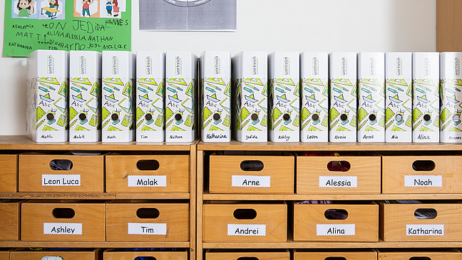 Auf einem Sideboard, in dem personalisierte Schubfächer der Schüler und Schülerinnen enthalten sind, stehen ebenfalls personalisierte wortreich Ordner.