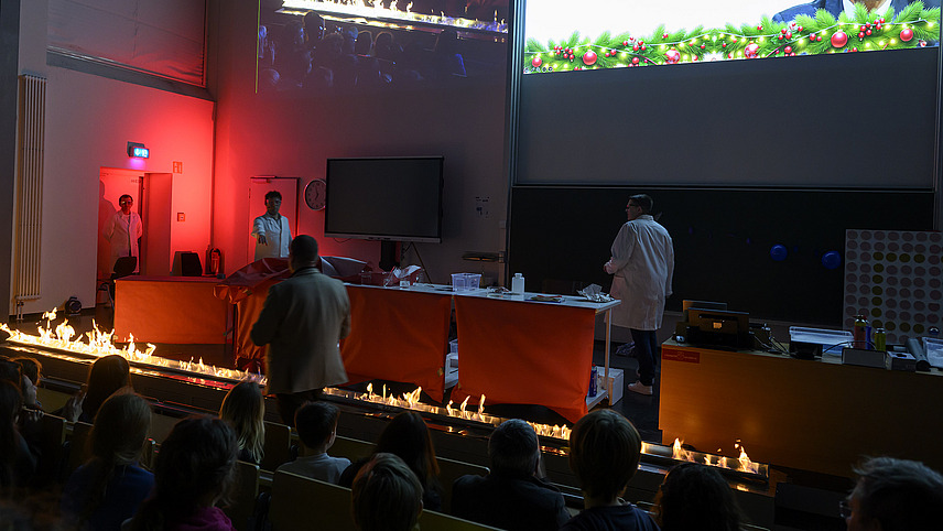 Personen in wei&szlig;en Kitteln unternehmen chemische Experimente in einem Vorlesungssaal, dabei entstehen Flammen