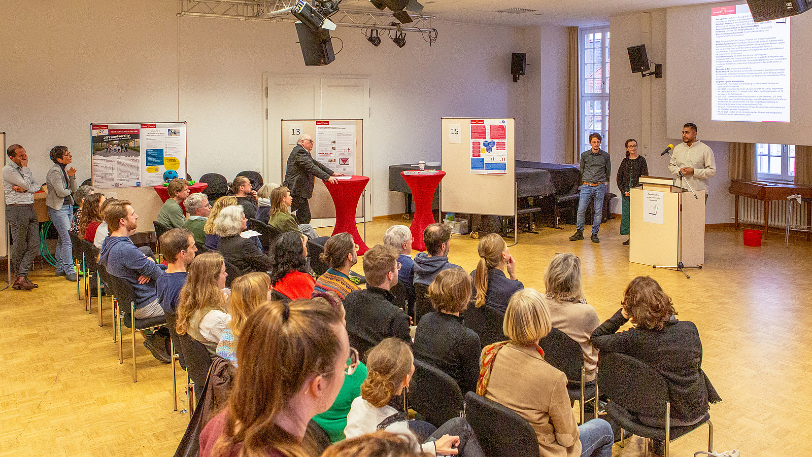 Viele Personen sitzen auf Stühlen und hören einem Vortragenden zu