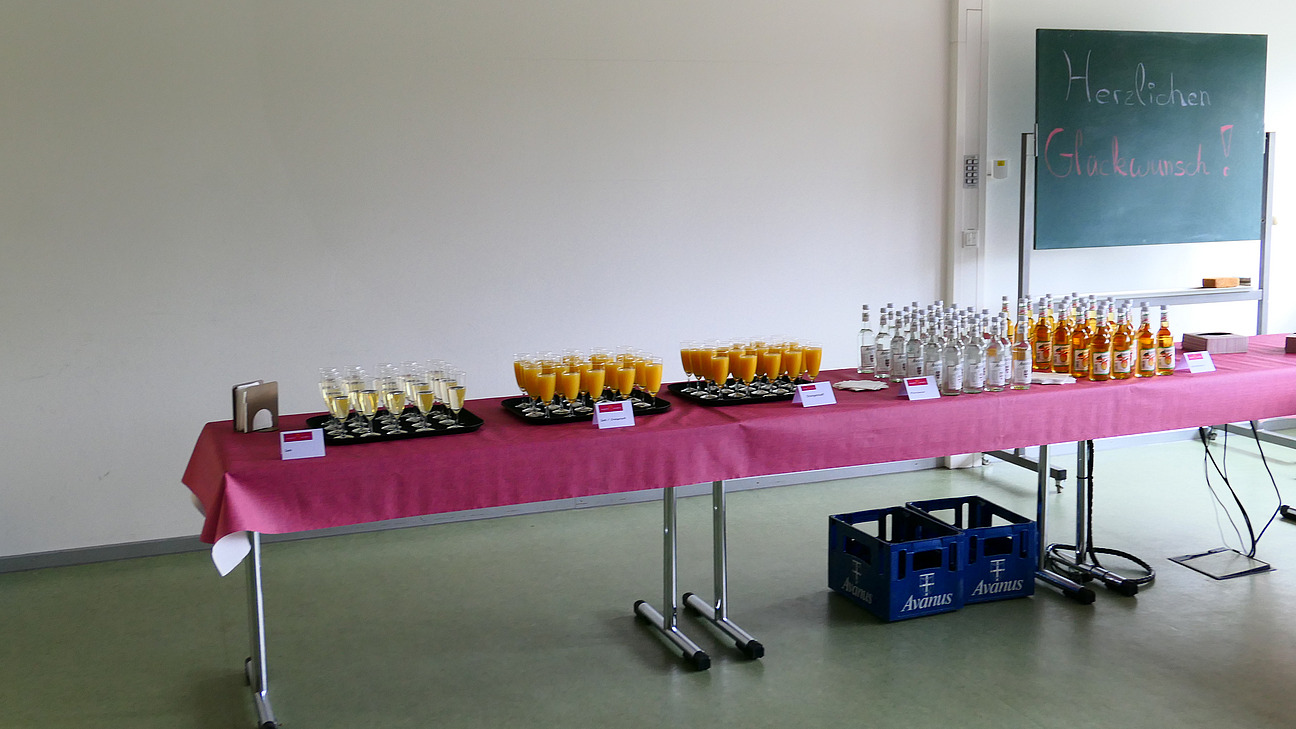 Gläser und Flaschen stehen auf einem Tisch vor einer Tafel. Auf der Tafel steht "Herzlichen Glückwunsch."