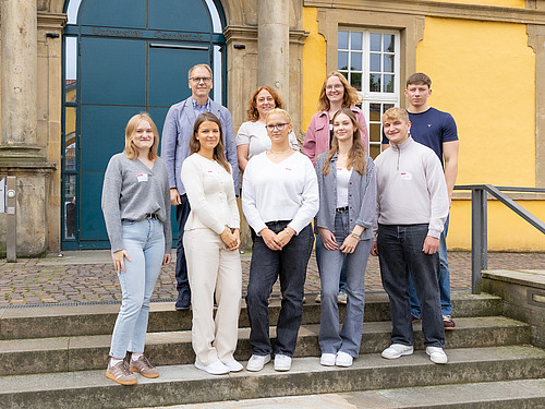 Eine Gruppe junger Menschen und zwei Menschen mittleren Alters stehen auf einer Steintreppe vor einem gro&szlig;en Geb&auml;ude
