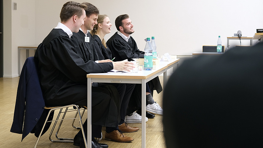 Team aus vier Personen in schwarzen Roben an Tischen sitzend und lachend