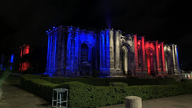 ruins of cartago at night, lit with red and blue spotlights. The stone architecture and gothic features are clearly visible. The ruins are surrounded by trimmed green hedges and a pathway.