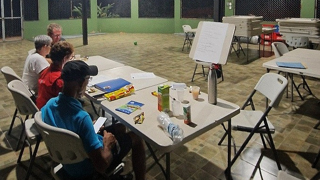 The photo shows a group of people gathered around a table in a shady outdoor area. They are taking part in an interview with Dennis Wilke. In front of them is a whiteboard with handwritten notes. On the table are documents, writing utensils, drinks and snacks.