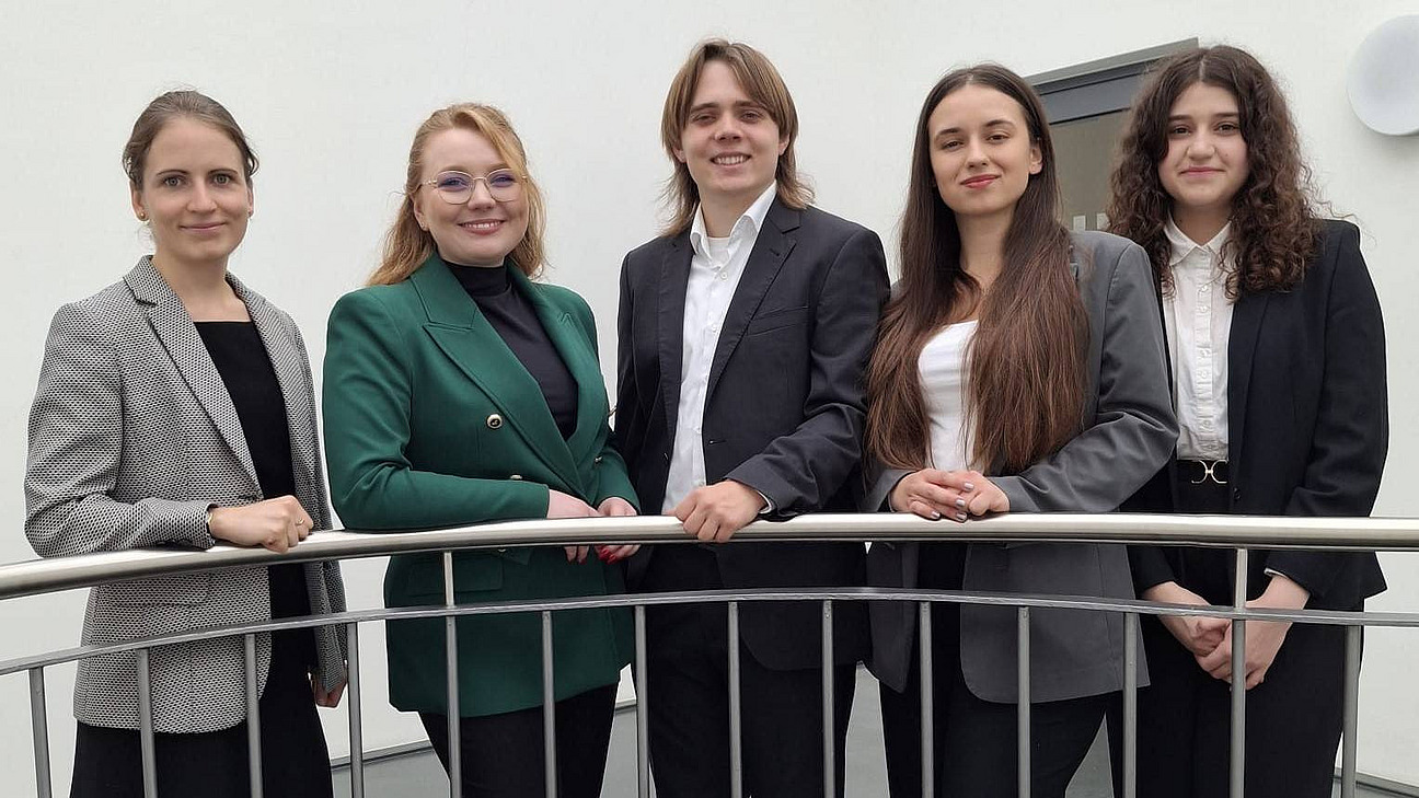 Vier Frauen und ein Mann in Anz&uuml;gen hinter einem halbhohen Metallgel&auml;nder stehend