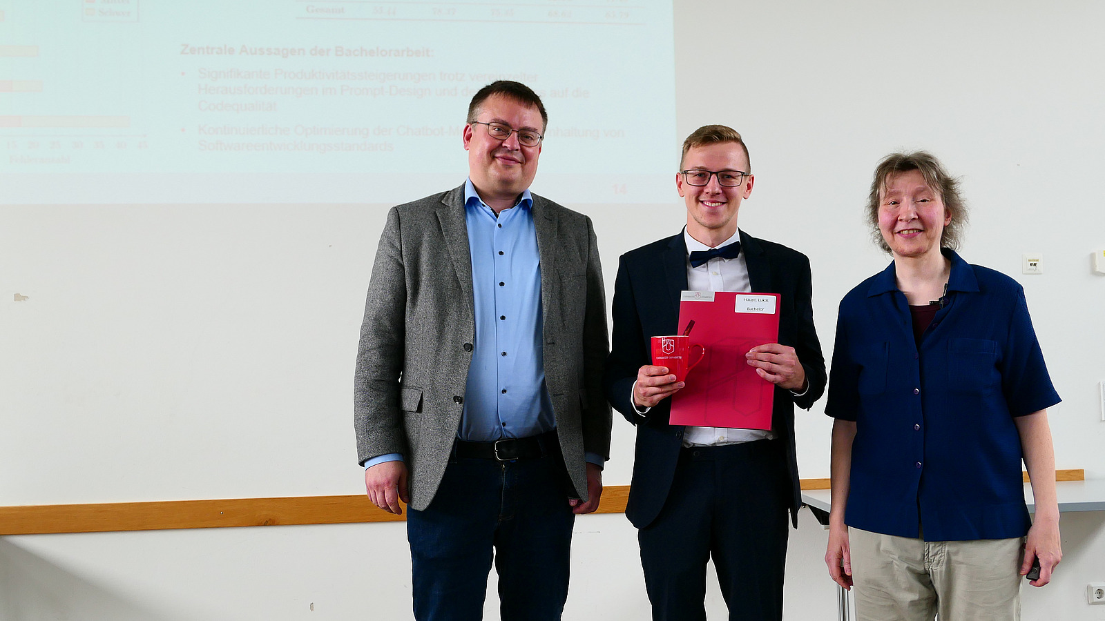 Ein Student mit Studiendekanin und Dekan stehen nebeneinander. Der Student hat eine Mappe in der Hand
