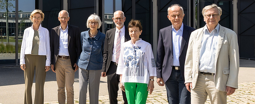 Eine Gruppe von sieben Personen in Business-Kleidung steht vor einem modernen Gebäude. Alle Personen lächeln in die Kamera