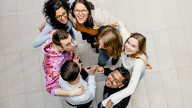 Ein Blick von Oben auf eine Gruppe von Studierenden dies sich im Kreis Umarmen und fröhlich nach Oben schauen.