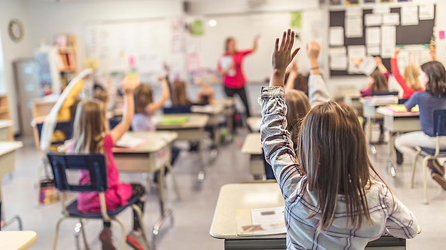 In a modern classroom, young pupils sit at their desks. Most of the children have raised their hands to participate in the lesson. In the background, the teacher stands pointing at one of the pupils. The wall behind her is decorated with learning materials, posters and notes. The atmosphere is lively and engaging, and the children are attentive and motivated.