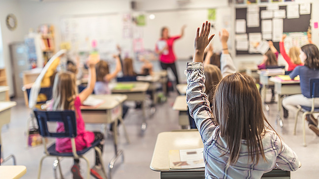 In a modern classroom, young pupils sit at their desks. Most of the children have raised their hands to participate in the lesson. In the background, the teacher stands pointing at one of the pupils. The wall behind her is decorated with learning materials, posters and notes. The atmosphere is lively and engaging, and the children are attentive and motivated.