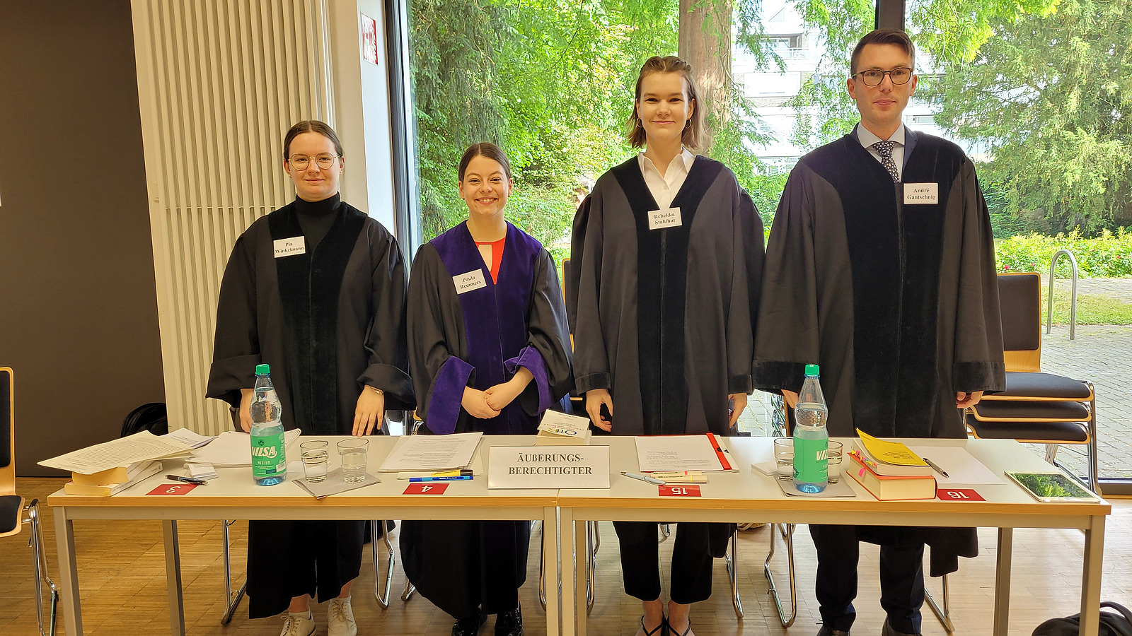 Ein Mann und drei Frauen in schwarzen Roben hinter einer Tischreihe stehend