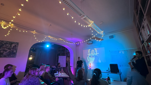 Ein Raum mit ged&auml;mpftem Licht, in dem mehrere Personen an Tischen sitzen und nach vorne schauen. An der Wand wird eine Pr&auml;sentation mit einem Beamer gezeigt. Eine Person steht vorne und spricht zur Gruppe.  Im Raum h&auml;ngen Lichterketten an der Decke, die eine gem&uuml;tliche Atmosph&auml;re schaffen. Das Licht ist in verschiedenen Farben gehalten, mit blauen und warmen T&ouml;nen. Auf den Tischen stehen Getr&auml;nke und pers&ouml;nliche Gegenst&auml;nde.