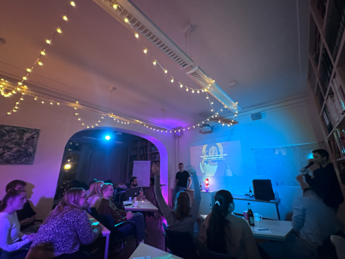 Ein Raum mit ged&auml;mpftem Licht, in dem mehrere Personen an Tischen sitzen und nach vorne schauen. An der Wand wird eine Pr&auml;sentation mit einem Beamer gezeigt. Eine Person steht vorne und spricht zur Gruppe.  Im Raum h&auml;ngen Lichterketten an der Decke, die eine gem&uuml;tliche Atmosph&auml;re schaffen. Das Licht ist in verschiedenen Farben gehalten, mit blauen und warmen T&ouml;nen. Auf den Tischen stehen Getr&auml;nke und pers&ouml;nliche Gegenst&auml;nde.